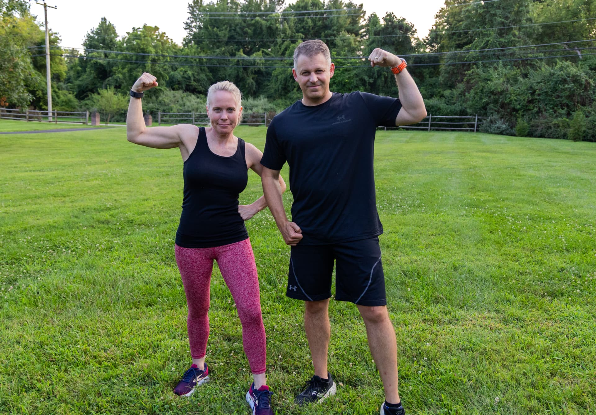 A man and a woman in athletic wear standing outdoors in a grassy field, flexing their arms